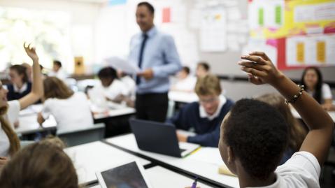 A classroom full of secondary school students working on tablets with the teacher walking between the desks in the background