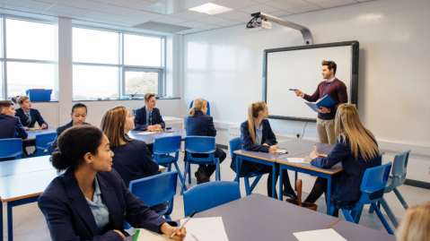A teacher stands in front of a smart board and projector in a secondary school classroom filled with students.